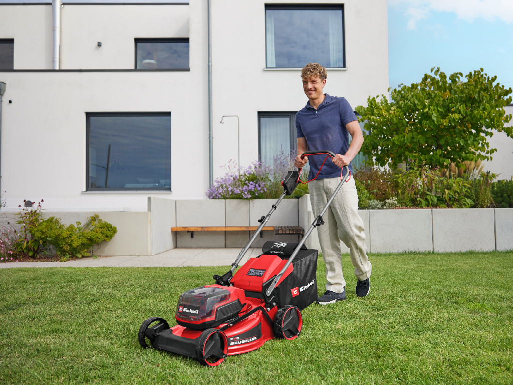 Auf dem Bild ist ein Mann mit rot-schwarz karierter Jacke zu sehen, der mit einem Einhell Akku-Laubbläser Laub aufwirbelt und wegbläst. 