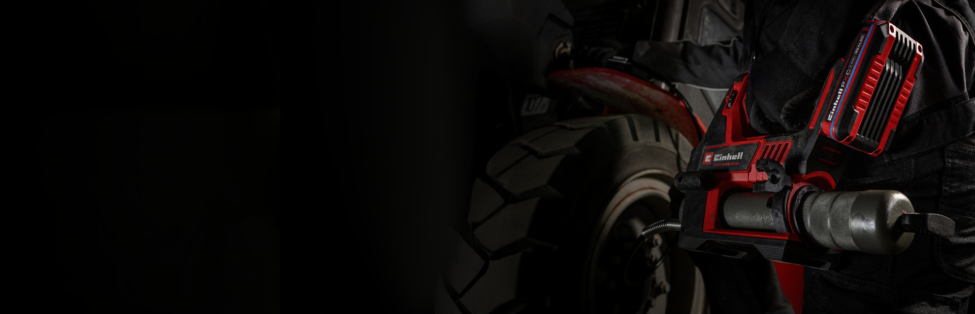 A person performs vehicle maintenance using a red cordless grease gun, wearing dark work clothing.