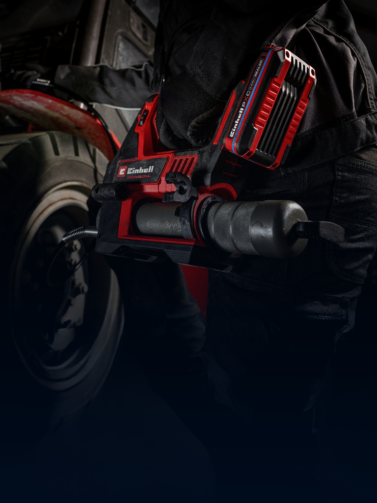 A person performs vehicle maintenance using a red cordless grease gun, wearing dark work clothing.
