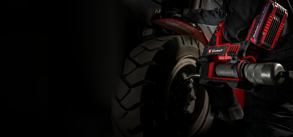 A person performs vehicle maintenance using a red cordless grease gun, wearing dark work clothing.