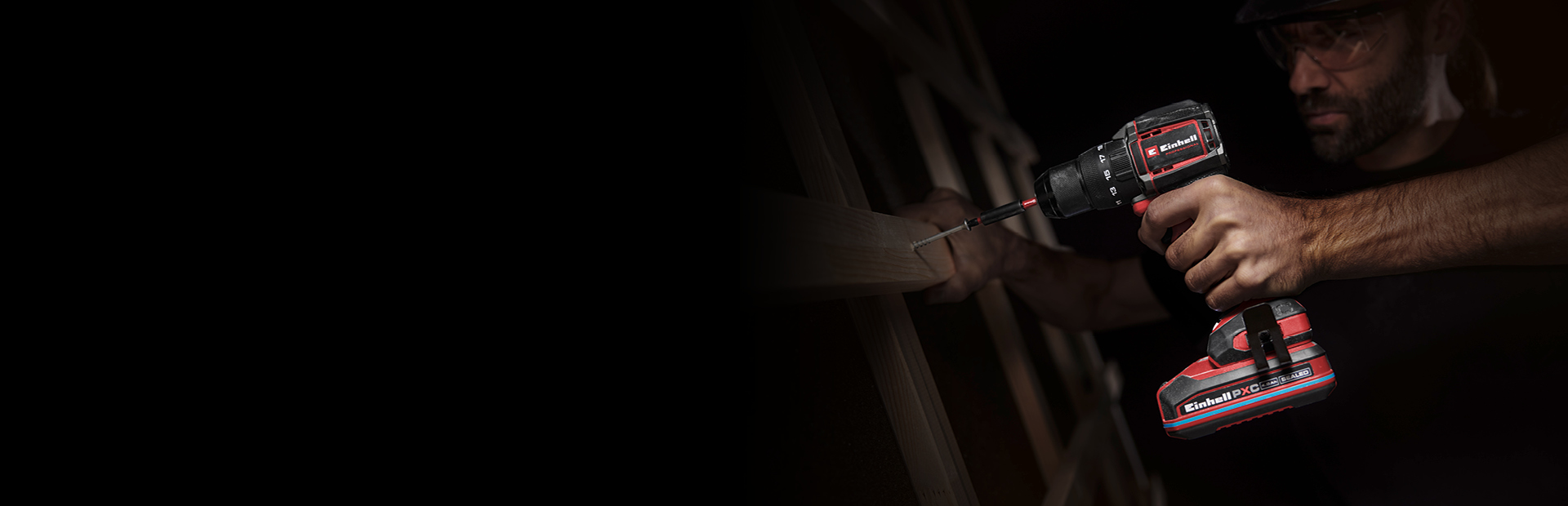 Ein Mann verwendet einen Akku-Schlagbohrschrauber, um eine Schraube in ein Stück Holz zu drehen. Die Szene ist dunkel, nur das Werkzeug und seine Hände sind im Vordergrund beleuchtet.