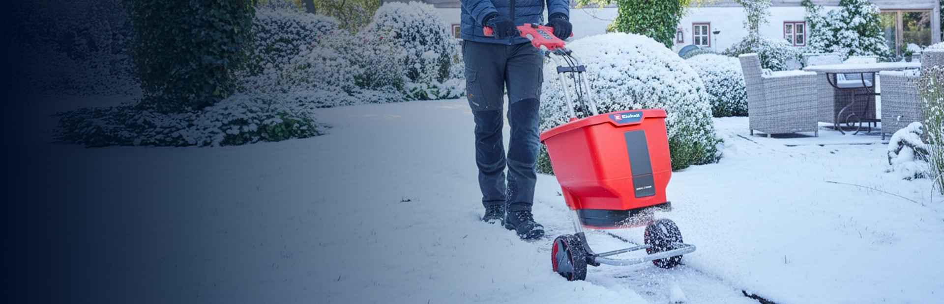 Eine Person benutzt einen roten Akku-Streuwagen auf einer schneebedeckten Gartenfläche in der Nähe eines Hauses und von Terrassenmöbeln. Der Boden, die Büsche und die Umgebung sind mit Schnee bedeckt.