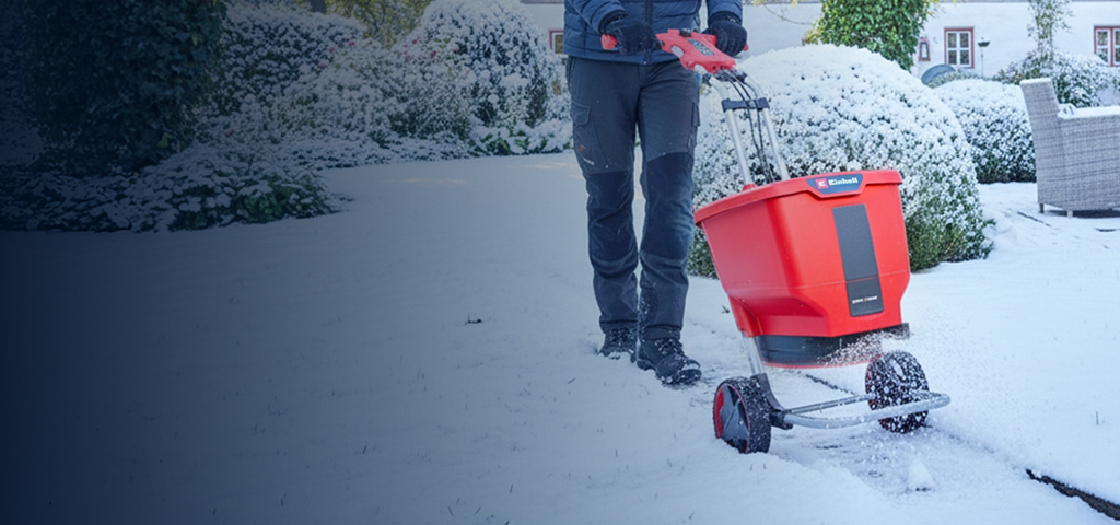 Eine Person benutzt einen roten Akku-Streuwagen auf einer schneebedeckten Gartenfläche in der Nähe eines Hauses und von Terrassenmöbeln. Der Boden, die Büsche und die Umgebung sind mit Schnee bedeckt.