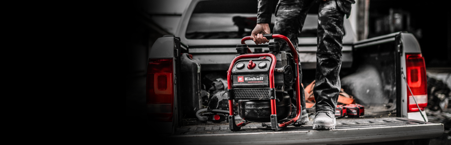Auf dem Bild steht ein tragbarer Einhell‑Kompressor auf der offenen Ladefläche eines Fahrzeugs und wird von einer Person am Griff gehalten. Die Szene wirkt wie eine Arbeitssituation, wobei der Kompressor farblich hervorsticht.