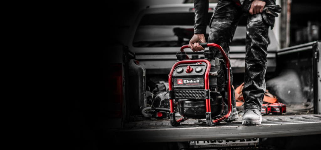 Auf dem Bild steht ein tragbarer Einhell‑Kompressor auf der offenen Ladefläche eines Fahrzeugs und wird von einer Person am Griff gehalten. Die Szene wirkt wie eine Arbeitssituation, wobei der Kompressor farblich hervorsticht.