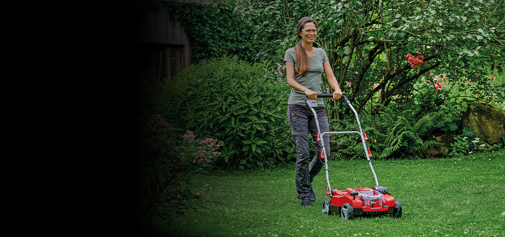 Die Aufnahme zeigt eine Person, die einen roten Rasenmäher über eine gepflegte, grüne Rasenfläche in einem üppigen Garten schiebt. Umgeben ist die Szene von dichtem Strauchwerk, Blumen und Bäumen.