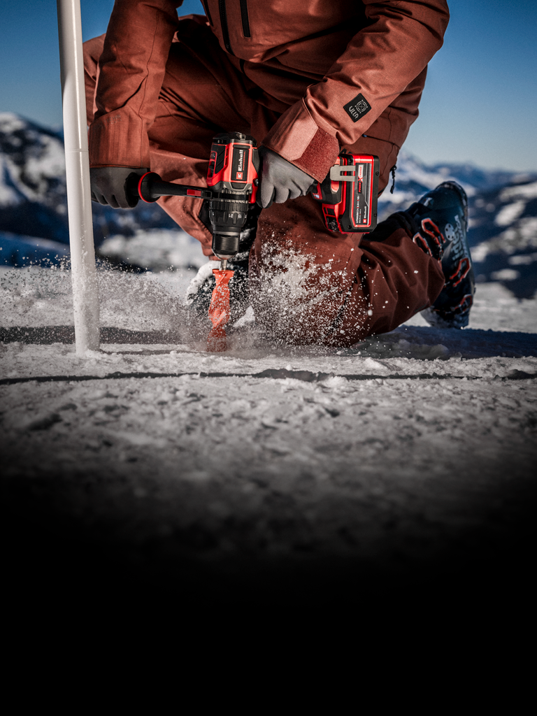 Eine Person in Winterkleidung bohrt mit einem kabellosen Elektrowerkzeug ein Loch in Eis oder verdichteten Schnee, wobei Schnee um den Bohrer herum aufwirbelt, in einer verschneiten Außenumgebung.