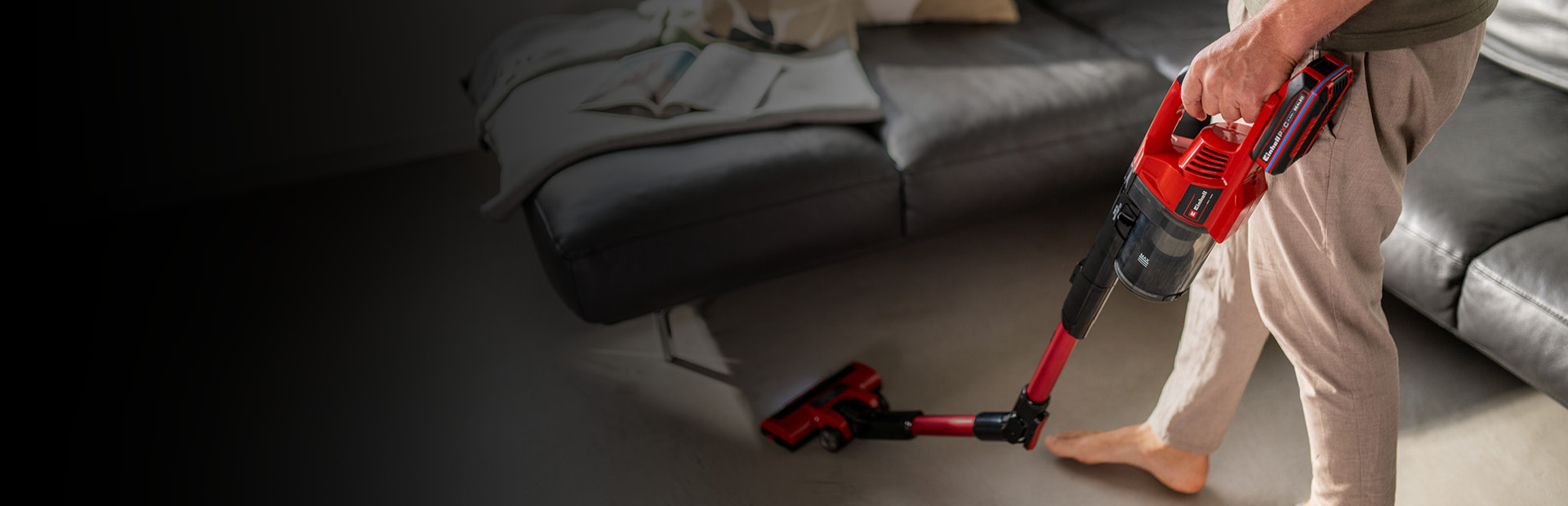 Eine Person saugt den Boden mit einem roten kabellosen Stielstaubsauger von Einhell. Zu sehen ist nur der untere Teil ihres Körpers, und sie steht barfuß in einem modernen Wohnzimmer. Im Hintergrund befindet sich ein graues Sofa mit einer locker darübergelegten Decke.
