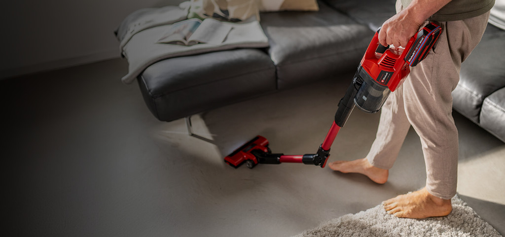Eine Person saugt den Boden mit einem roten kabellosen Stielstaubsauger von Einhell. Zu sehen ist nur der untere Teil ihres Körpers, und sie steht barfuß in einem modernen Wohnzimmer. Im Hintergrund befindet sich ein graues Sofa mit einer locker darübergelegten Decke.