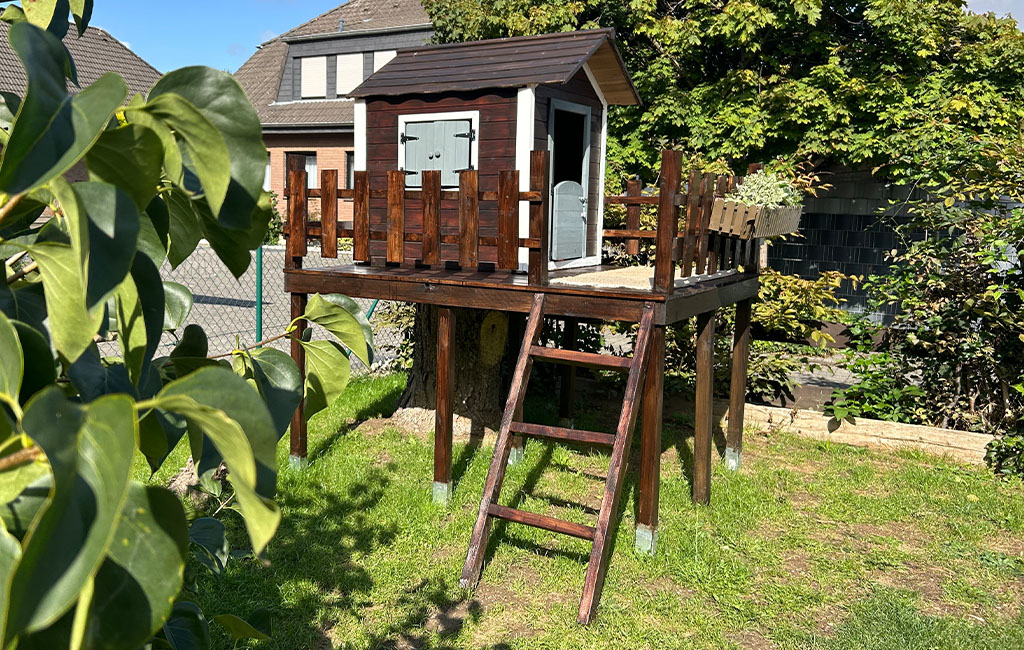 Das fertige Spielhaus mit dunkel lasiertem Holzgeländer steht erhöht auf Stelzen im Garten.