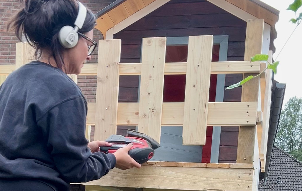 Eine Frau schleift das Holzgeländer des Spielhauses mit einem Handschleifer ab.