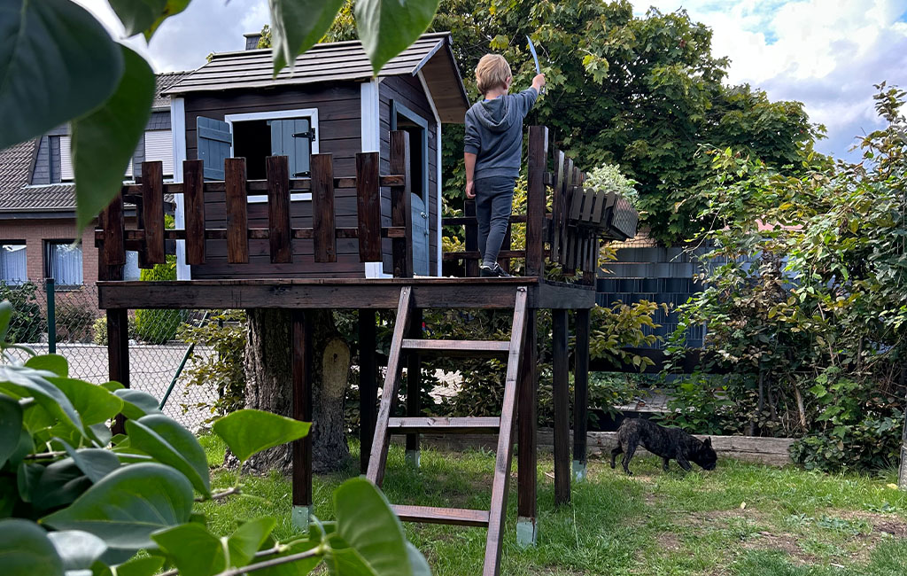 Ein Junge steht auf einem erhöhten Spielhaus mit Holzgeländer und hält ein Spielschwert in der Hand. Ein Hund läuft unten auf dem Rasen.