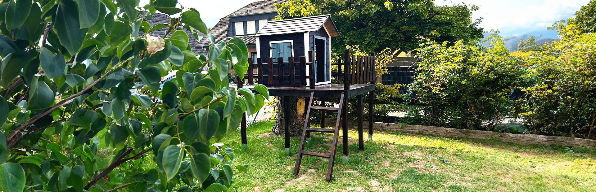 Das fertige Spielhaus mit Holzgeländer steht erhöht auf Stelzen im Garten, neben einem Baumstumpf.