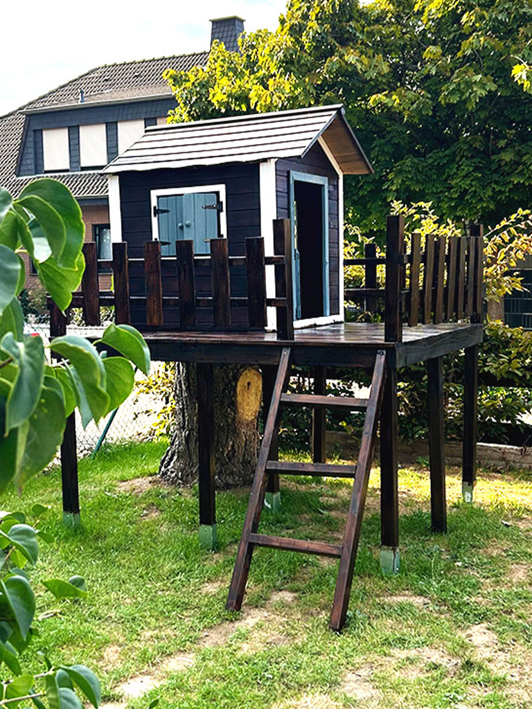 Das fertige Spielhaus mit Holzgeländer steht erhöht auf Stelzen im Garten, neben einem Baumstumpf.