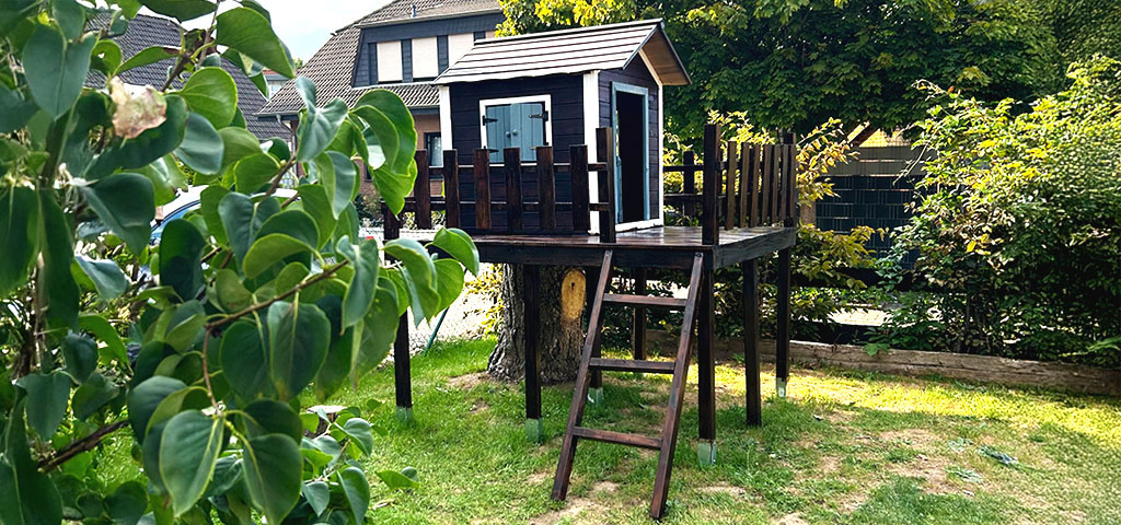 Das fertige Spielhaus mit Holzgeländer steht erhöht auf Stelzen im Garten, neben einem Baumstumpf.