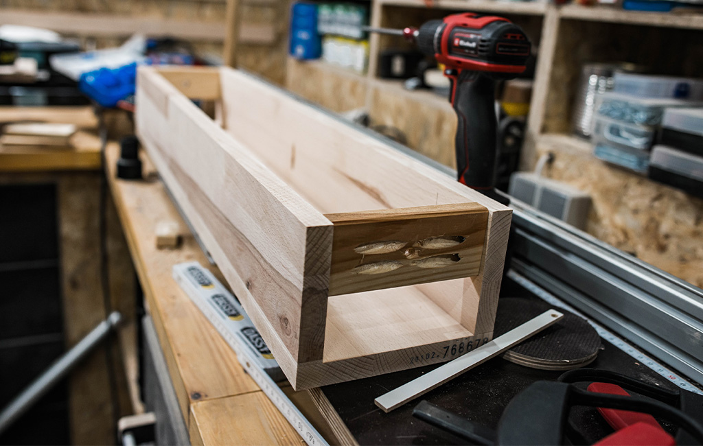 Zusammengebauter Holzkasten mit sichtbaren Schraubverbindungen auf Werkbank, dahinter ein Einhell Akkuschrauber.