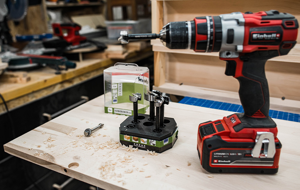 Ein Einhell Akkuschrauber mit Forstnerbohrern und Verpackung der Marke kwb auf einer Holzplatte in der Werkstatt.