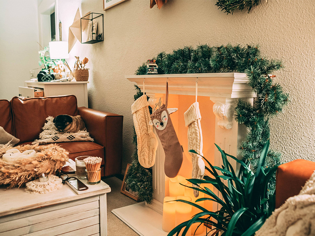 Weihnachtlich geschmücktes Wohnzimmer mit Kaminattrappe, Strümpfen und Lichterdekoration; ein Hund liegt auf dem Sofa.