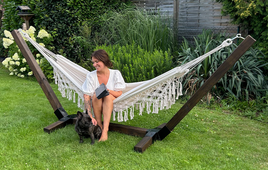 Eine Frau sitzt in der Hängematte mit dunklem Holzgestell im Garten und streichelt einen schwarzen Hund.
