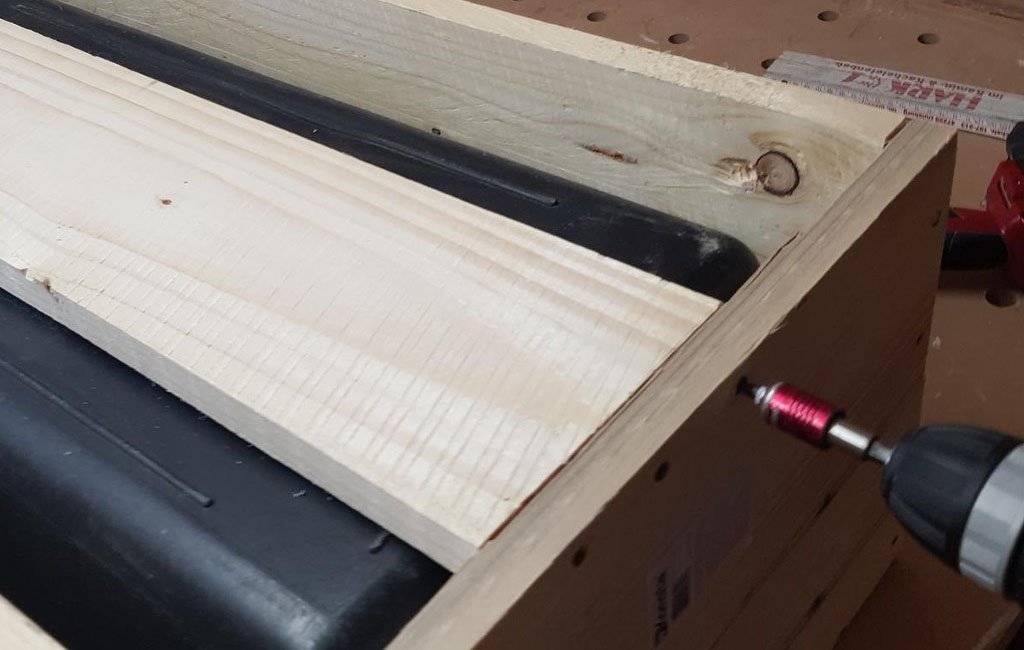 Eine Holzlatte wird mit einer Schraube an einen Holzkasten geschraubt, daneben ist die schwarze Kunststoffbox zu sehen.