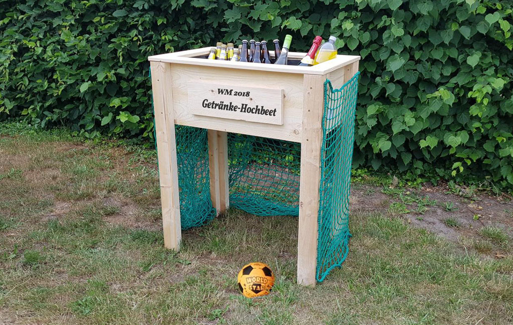 Ein Hochbeet mit Flaschen als Getränkebar, an den Seiten grüne Netze. Text auf dem Beet: „WM 2018 Getränke-Hochbeet“.