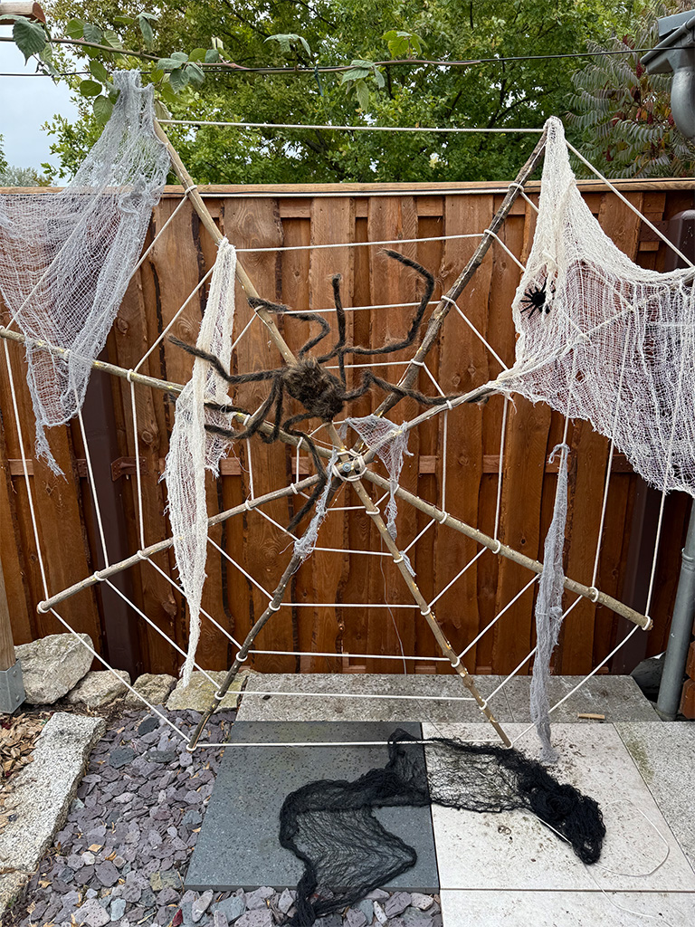 Large spider web made of ropes with white cloth and two artificial spiders, mounted on a wooden frame in a garden.