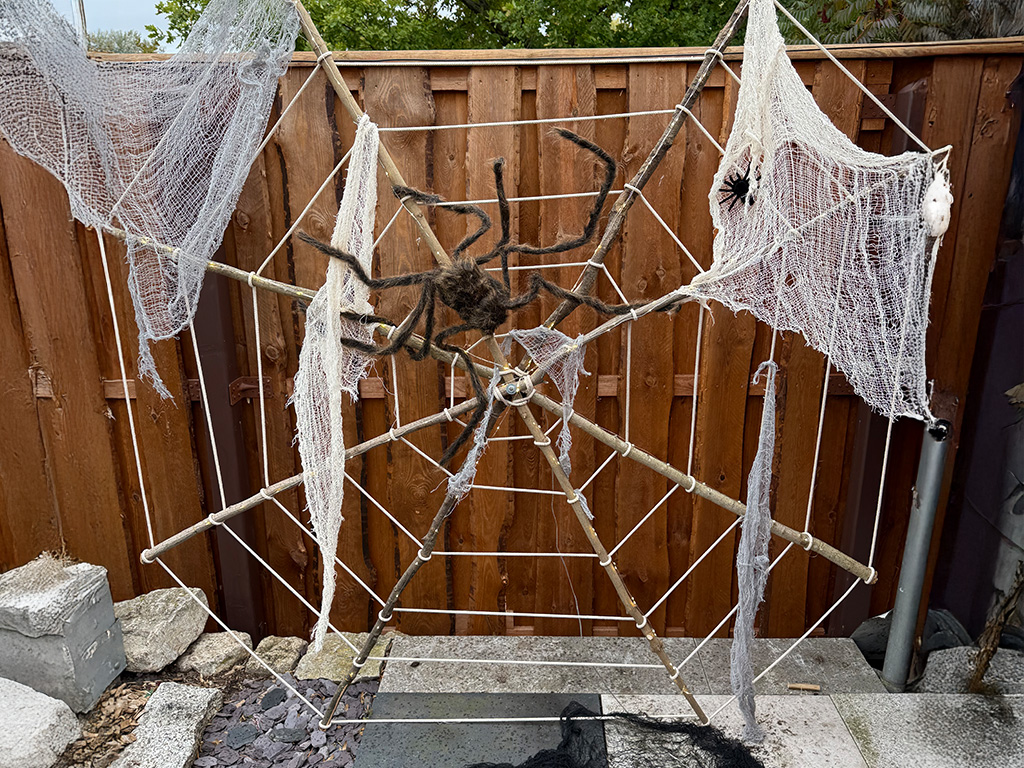 Large spider web made of ropes with white cloth and two artificial spiders, mounted on a wooden frame in a garden.